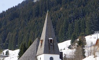 A charming church with a pointed tower in a snow-covered landscape. In the background, green mountains are visible. | © Ev. Kirche Kleinwalsertal