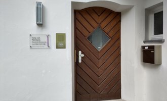 A brown front door with a diamond-shaped window. Next to the door is a mailbox and a sign with information. | © Ev. Kirche Kleinwalsertal