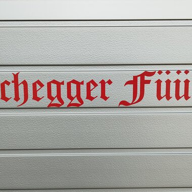 A sign with the inscription "Hirschegger Feuerwehr" in red letters. The letters are designed in a decorative font. | © Kleinwalsertal Tourismus