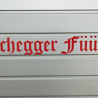 A sign with the inscription "Hirschegger Feuerwehr" in red letters. The letters are designed in a decorative font. | © Kleinwalsertal Tourismus