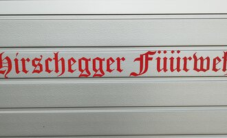 A sign with the inscription "Hirschegger Feuerwehr" in red letters. The letters are designed in a decorative font. | © Kleinwalsertal Tourismus
