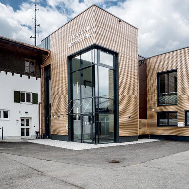 A modern building with a wooden façade and large glass windows. It is located in a rural area with other traditional buildings. | © Feuerwehr Riezlern | Gemeinde Mittelberg