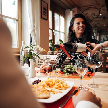 A woman smiles in a restaurant while her wine is being poured. On the table, there is also a plate of fries and a portion of salad.