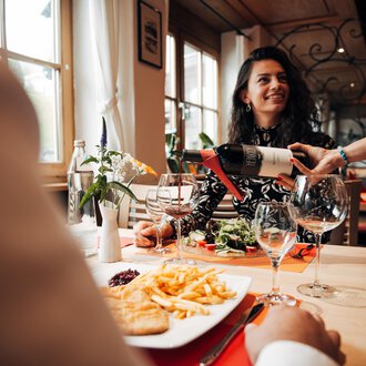 A woman smiles in a restaurant while her wine is being poured. On the table, there is also a plate of fries and a portion of salad.