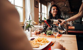 A woman smiles in a restaurant while her wine is being poured. On the table, there is also a plate of fries and a portion of salad.