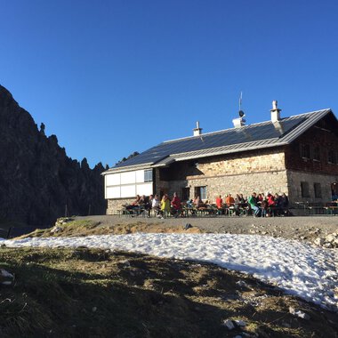 Eine Berghütte in den Alpen umgeben von schneebedecktem Boden. Viele Menschen sitzen draußen und genießen die Sonne unter blauen Himmel. | © Kleinwalsertal Tourismus