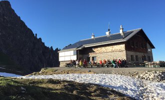 A mountain hut in the Alps surrounded by snow-covered ground. Many people are sitting outside enjoying the sun under a blue sky. | © Kleinwalsertal Tourismus