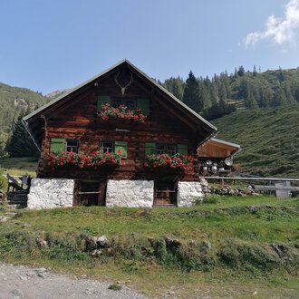 © Fluchtalpe | N. Lughammer Ein rustikales Holzhaus umgeben von grünen Wiesen und Bergen. Blumenkästen mit bunten Blumen schmücken die Fenster. | © Fluchtalpe | N. Lughammer
