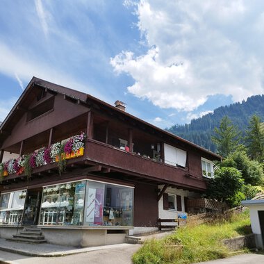 Ein charmantes Haus mit einem blühenden Balkon in den Bergen. Die Umgebung ist grün und die Wolkenhimmel sorgt für eine schöne Atmosphäre. | © Kleinwalsertal Tourismus | N. Lughammer