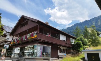 A charming house with a blooming balcony in the mountains. The surroundings are green, and the cloudy sky creates a beautiful atmosphere. | © Kleinwalsertal Tourismus | N. Lughammer
