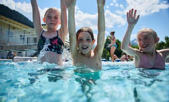 Three cheerful children are splashing in the swimming pool and having a lot of fun. They lift their hands in the air and enjoy the sun. | © Kleinwalsertal Tourismus | Oliver Farys