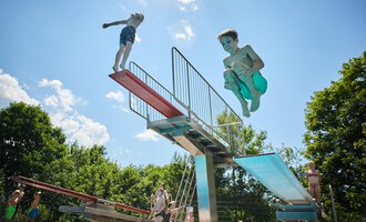 Children jump from a diving tower into the water. It's a sunny day with a blue sky and green trees in the background. | © Kleinwalsertal Tourismus | Oliver Farys