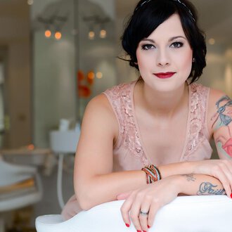 © Fräulein Sugar Coiffeur | Sandra Bromm A woman with tattoos is sitting smiling in a modern room. She is wearing an elegant pink dress and has a chic look. | © Fräulein Sugar Coiffeur | Sandra Bromm
