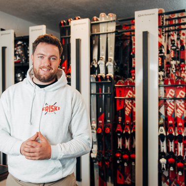 A man is smiling in a ski rental shop with many skis in the background. He is wearing a white hoodie and looks inviting and friendly. | © Friski Marcellus Fritz | Stefan Klauser