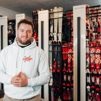 A man is smiling in a ski rental shop with many skis in the background. He is wearing a white hoodie and looks inviting and friendly. | © Friski Marcellus Fritz | Stefan Klauser