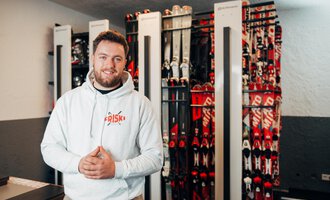 A man is smiling in a ski rental shop with many skis in the background. He is wearing a white hoodie and looks inviting and friendly. | © Friski Marcellus Fritz | Stefan Klauser