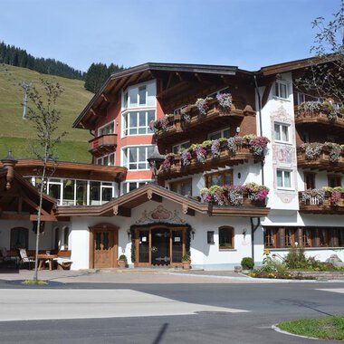 A charming building in alpine style with many balconies and flowers. In the background, green hills and a clear sky can be seen. | © Hotel Alpenstüble Appartements | Manuel Riezler
