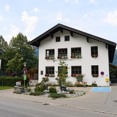Ein charmantes Gemeindegebäude mit blühenden Blumenkästen. Im Hintergrund sind weitere Gebäude und Bäume zu sehen. | © Kleinwalsertal Tourismus | Oliver Farys