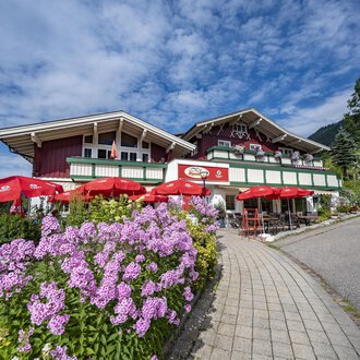 A picturesque alpine house with red sunshades and colorful flowers. The sky is blue with some clouds. | © Gasthof Alpenblick | Theodor Pinn