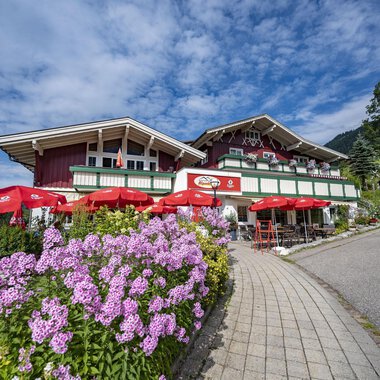 A picturesque alpine house with red sunshades and colorful flowers. The sky is blue with some clouds. | © Gasthof Alpenblick | Theodor Pinn