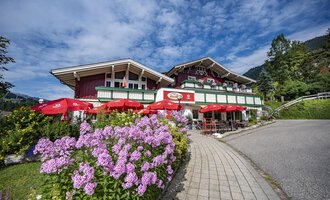 Ein malerisches alpenländisches Haus mit roten Sonnenschirmen und bunten Blumen. Der Himmel ist blau mit einigen Wolken. | © Gasthof Alpenblick | Theodor Pinn
