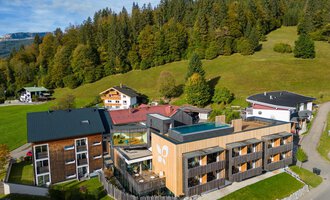 Ein modernes Gebäude im Grünen, umgeben von Bäumen und Hügeln. Die Architektur kombiniert Holz und Glas und vermittelt eine einladende Atmosphäre.