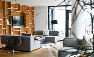 Ein modernes Wohnzimmer mit grauen Sofas und einem großen Fernseher. Große Fenster sorgen für viel Licht und einen offenen Raum. | © Katharina Wohlgenannt