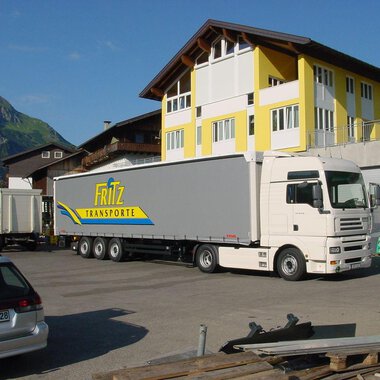 Ein Lkw von Fritz Transporte steht auf einem Parkplatz vor einem großen Gebäude. Im Hintergrund sind grüne Berge zu sehen. | © Getränke Service Fritz | Helmut Fritz
