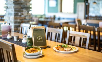 Ein gemütliches Restaurant mit Holzstühlen und einem sauberen Tisch. Auf dem Tisch liegt eine Speisekarte, daneben befinden sich ein Teller mit Fleisch und Gemüse sowie eine Schüssel mit Reis. | © OBERSTDORF·KLEINWALSERTAL BERGBAHNEN
