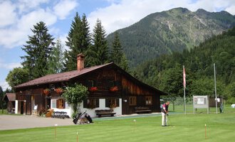Ein traditionelles Holzhaus umgeben von Bergen und Bäumen. Im Vordergrund sieht man einen gepflegten Golfplatz. | © Golfclub Oberstdorf | ©kunst.joachimweiler.de