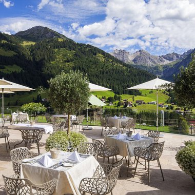 Ein stilvolles Restaurant im Freien mit Blick auf die Berge. Es gibt elegante Tische mit Sonnenschirmen und grüne Pflanzen. | © Hallers Geniesserhotel | Dietmar Walser