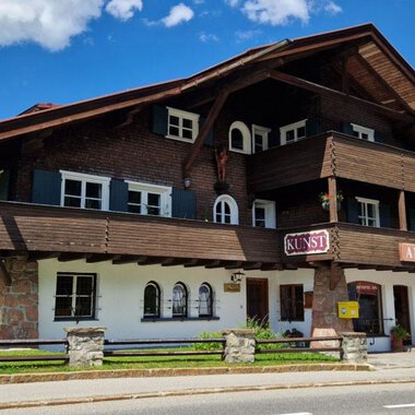 Ein charmantes, traditionelles Chalet aus Holz mit mehreren Balkonen und Fenstern. Der Himmel ist blau und voller Wolken, während der grüne Rasen das Gebäude umgibt. | © Antik und Kunst im Malerhäusl Herbert Stumvoll