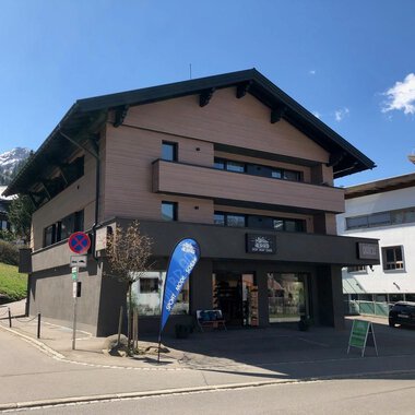 A modern building at a street corner with a shop on the ground floor. In the background, mountains and other buildings can be seen. | © Sport Hilbrand GmbH