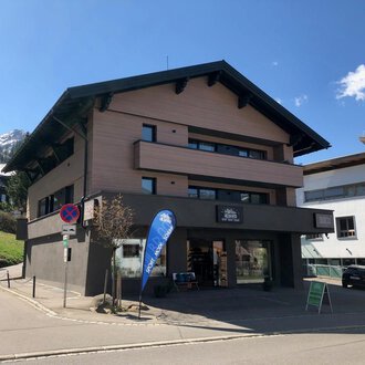 Ein modernes Gebäude an einer Straßenecke mit einem Geschäft im Erdgeschoss. Im Hintergrund sind Berge und andere Gebäude zu sehen. | © Sport Hilbrand GmbH