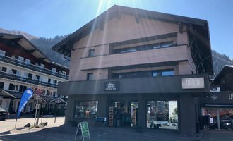A modern building with a shop on the ground floor. The sun is shining on the facade, and in the background, there are more houses and mountains visible. | © Sport Hilbrand OG