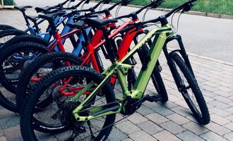 Several mountain bikes in different colors are standing on a gravel path. In the background, a wooden house and a green meadow can be seen. | © Sport Hilbrand GmbH