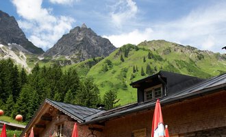 Ein gemütliches Restaurant in den Bergen mit Terrassen und roten Sonnenschirmen. Im Hintergrund sind majestätische Berge und ein blauer Himmel zu sehen. | © Hintere Gemstelhütte | Frank Drechsel