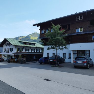 A quiet village with traditional houses and cars on the street. Green hills are visible in the background. | © Kleinwalsertal Tourismus | Nicole Lughammer