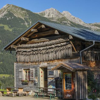 Ein traditionelles Holzhaus in den Alpen mit einem grünen Landschaftshintergrund. Die Berge sind klar und sonnig sichtbar. | © Hoflaada | Dagmar Hilbrand