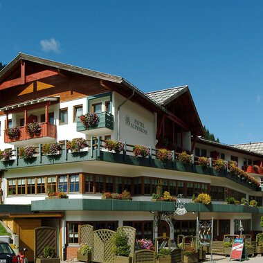 Ein charmantes Hotel im alpinen Stil mit einem welligen Dach und vielen Balkonen. Umgeben von grünen Hügeln und blauer Himmel. | © IFA Hotel Alpenrose
