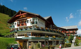 Ein charmantes Hotel im alpinen Stil mit einem welligen Dach und vielen Balkonen. Umgeben von grünen Hügeln und blauer Himmel. | © IFA Hotel Alpenrose