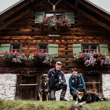 A rustic wooden house with blooming flower boxes. A couple with two dogs poses in front of it. | © Hundewelt Kleinwalsertal | Stefan Lindbauer