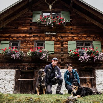 A rustic wooden house with blooming flower boxes. A couple with two dogs poses in front of it. | © Hundewelt Kleinwalsertal | Stefan Lindbauer