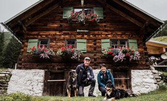 A rustic wooden house with blooming flower boxes. A couple with two dogs poses in front of it. | © Hundewelt Kleinwalsertal | Stefan Lindbauer