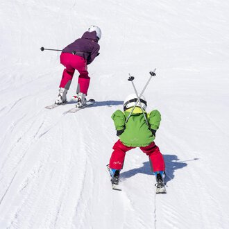 Zwei Kinder fahren Ski auf einer verschneiten Piste. Das eine Kind trägt einen grünen Anzug und das andere einen lila Anzug. | © Kleinwalsertal Tourismus | Dominik Berchtold