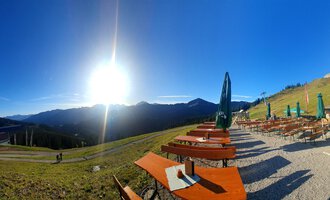 Eine schöne Berglandschaft mit Sonne, die über den Gipfeln scheint. Im Vordergrund stehen Holzsitze und Tische mit grünen Sonnenschirmen. | © Ifenhütte | Petra Diener