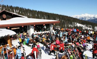 Eine belebte Skihütte in den Bergen, umgeben von Schnee und Bäumen. Viele Menschen genießen die Sonne und die winterliche Atmosphäre. | © Ifenhütte | Petra Diener
