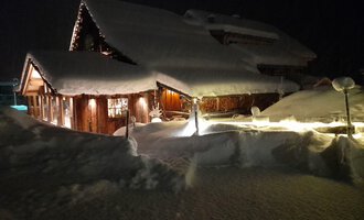 A cozy house in the snow at night, illuminated by warm light. The snow covers the surroundings and creates a wintry atmosphere.