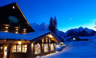 The cozy iKuh shop in an old farmhouse in the snow under a clear blue sky. The mountains in the background are softly illuminated as evening approaches.