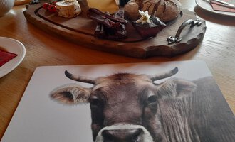 A rustic wooden board with various types of cheese, sausage, and fresh bread. The table is cozily set, and in the foreground, there is a placemat with a picture of a cow.
